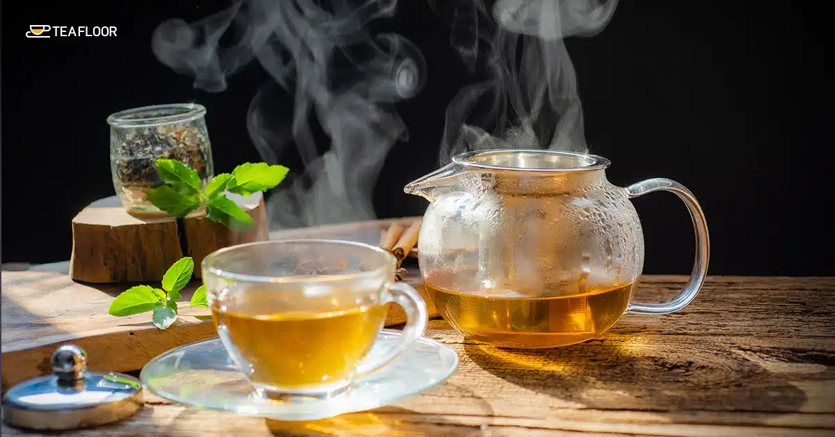 freshly brewed hot beverage in a transparent cup next to a steaming glass teapot on a rustic wooden table with fresh green herbs